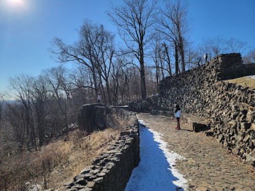 A stone pathway winds through a winter landscape with bare trees and patches of snow under a clear blue sky.