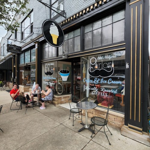 A quaint ice cream shop with outdoor seating, featuring a sign with a soft serve cone and large windows displaying treats.