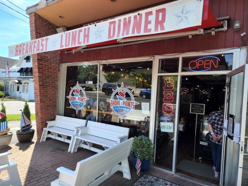 Exterior of a restaurant with a sign reading "Breakfast Lunch Dinner" and "OPEN," featuring BBQ-themed decor and seating.