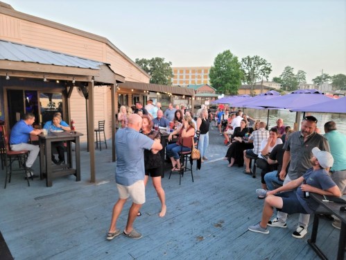 A lively outdoor gathering at a waterfront venue with people socializing, dancing, and enjoying the evening atmosphere.