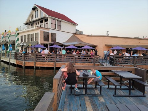 A waterfront scene with people dining at outdoor tables, a child playing, and a lively restaurant in the background.