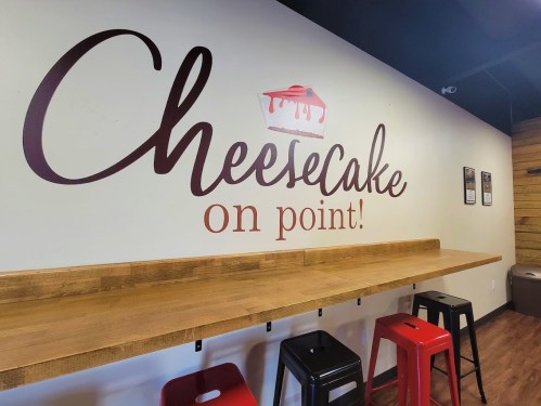 A wall with the text "Cheesecake on point!" above a wooden counter and colorful stools in a cozy setting.
