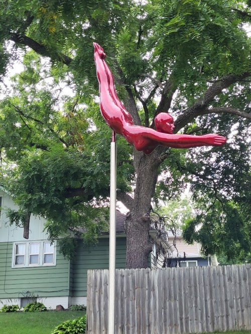A bright red sculpture of a diver leaping from a pole, set against a backdrop of trees and a house.