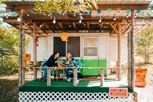 Two women sit on a porch of a cozy camper, enjoying drinks and conversation in a vibrant outdoor setting.