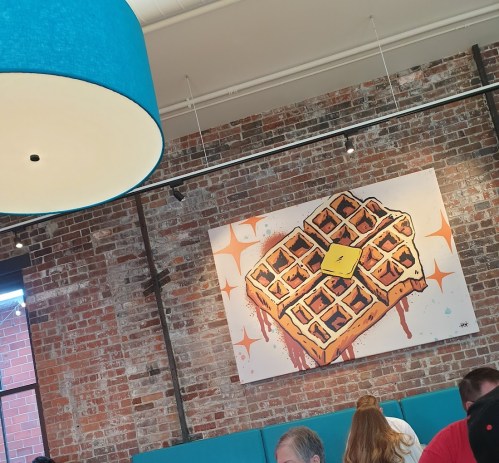 A colorful waffle mural on a brick wall in a cozy café, with patrons seated nearby.