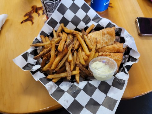 A basket of golden fries and two pieces of fried fish, served with a small cup of dipping sauce.