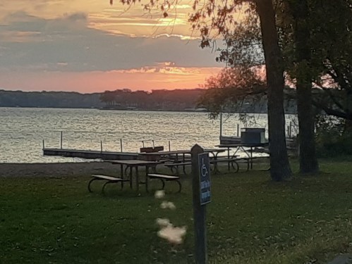 Sunset over a lake with picnic tables and a dock, framed by trees and grassy shoreline.