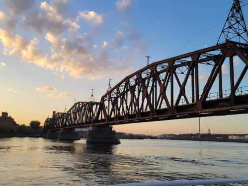 A rusted bridge spans a calm river at sunset, with clouds and a city skyline in the background.