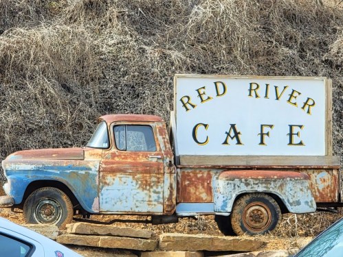 An old, rusty pickup truck with a sign reading "Red River Cafe" parked on a rocky surface.