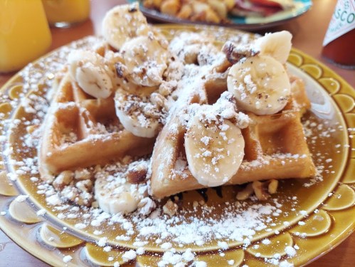 A plate of waffles topped with sliced bananas, crushed nuts, and powdered sugar, served with a side of syrup.
