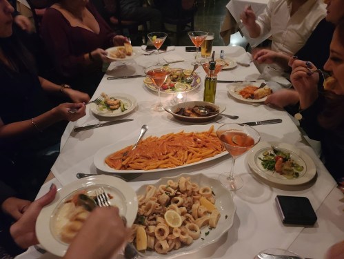 A table filled with various dishes, including pasta, seafood, salads, and drinks, with people enjoying their meal.