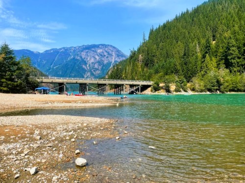 A serene river scene with a bridge, surrounded by lush green mountains and a clear blue sky.