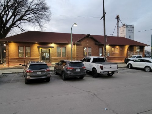 A brick building with a red roof, surrounded by parked cars, in a parking lot during dusk.