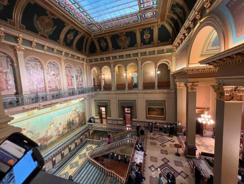 A grand interior of a historic building featuring ornate architecture, murals, and a sweeping staircase.