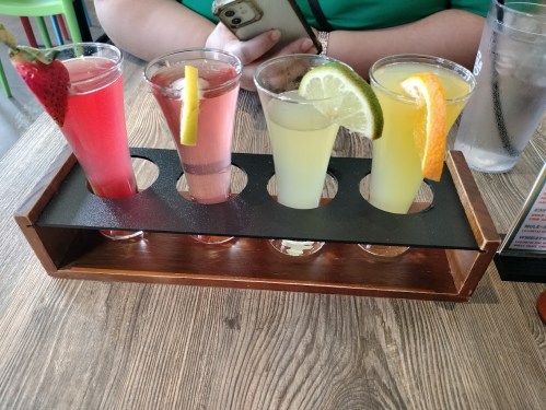 Four colorful shot glasses on a wooden tray, featuring drinks with strawberry, lemon, lime, and orange garnishes.