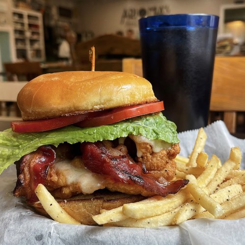A close-up of a chicken sandwich with bacon, lettuce, and tomato, served with fries and a drink in the background.