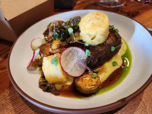 A plate of braised meat with crispy potatoes, greens, and garnishes, served on a creamy base with a drizzle of sauce.