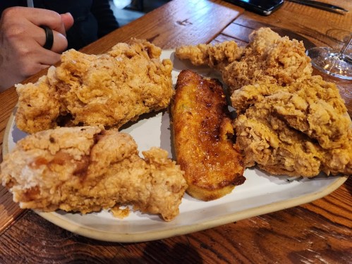 A plate of crispy fried chicken pieces alongside a slice of golden-brown cornbread on a wooden table.