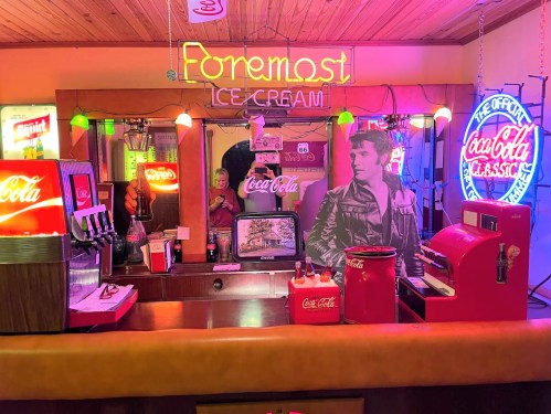 A retro ice cream shop with neon signs, vintage Coca-Cola decor, and a large photo of a man in a leather jacket.