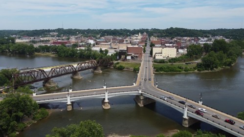 Aerial view of a city with a bridge over a river, surrounded by greenery and buildings in the background.