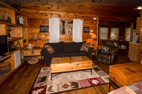 Cozy cabin living room with wooden walls, a black sofa, a coffee table, and warm lighting. Decor includes bear-themed rug.