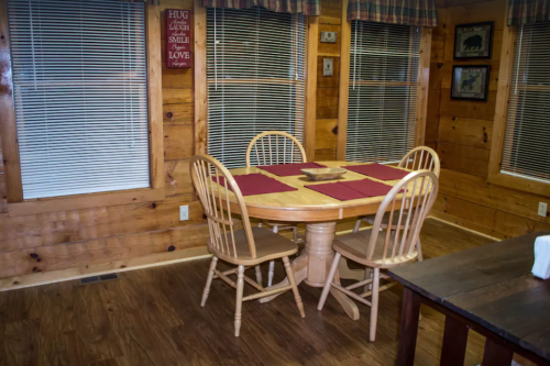 Cozy wooden dining area with a round table, four chairs, and large windows with blinds. Warm, inviting atmosphere.