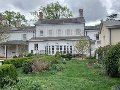 A large, historic white house with a garden, surrounded by greenery and cloudy skies.
