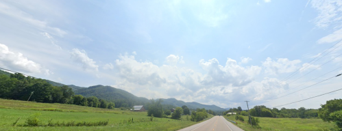 A scenic view of a rural road lined with green fields and mountains under a partly cloudy sky.