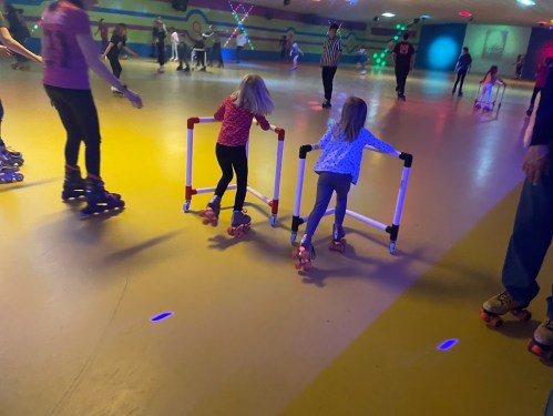 Two young girls on roller skates use support frames while skating in a colorful roller rink with others around them.