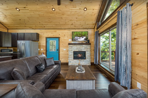 Cozy cabin interior with wooden walls, a stone fireplace, modern kitchen, and large windows overlooking nature.