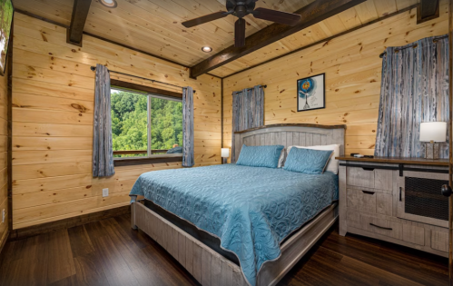 Cozy bedroom with wooden walls, a queen bed with a blue quilt, and a window overlooking greenery.