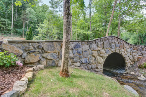 A stone bridge with a curved design over a small stream, surrounded by lush greenery and trees.