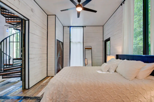 A cozy bedroom with a spiral staircase, large windows, and a bed with white bedding against a wooden floor.