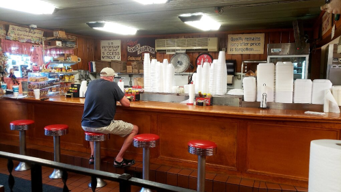 A diner with red stools and a counter, featuring stacks of white cups and condiments, and a lone customer sitting.