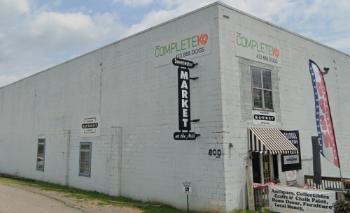 A white building with signs for "Complete K9" and "Market at the Mill," featuring a striped awning and outdoor displays.