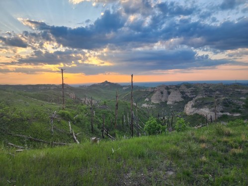 A scenic landscape at sunset, featuring rolling hills, green vegetation, and dramatic clouds in the sky.