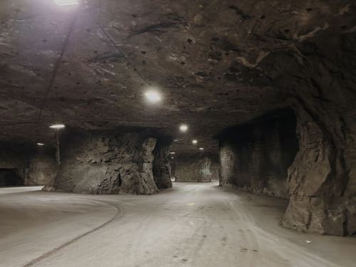 A dimly lit underground tunnel with rocky walls and a curved path, illuminated by overhead lights.