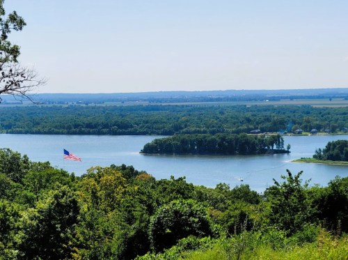 A scenic view of a lake with an island, surrounded by lush greenery and a distant American flag.
