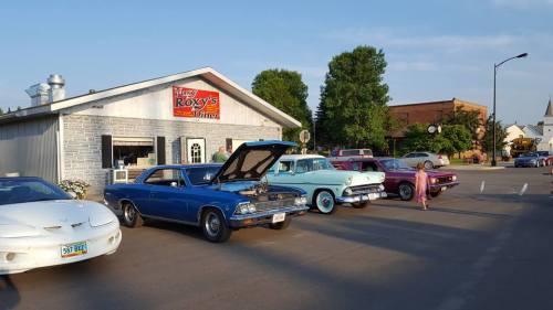 A row of classic cars parked outside Roxy's Diner on a sunny day, with a blue sky and trees in the background.