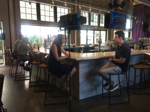 A woman interviews a man at a bar, with patrons in the background and a camera set up for recording.