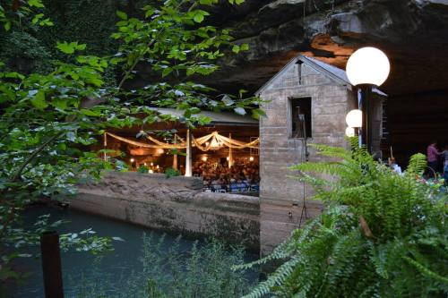 A rustic venue inside a cave, featuring a stone structure, greenery, and soft lighting over a tranquil waterway.