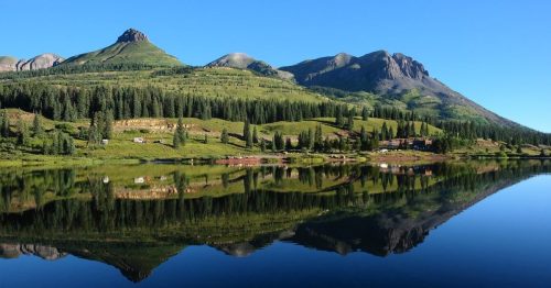A serene mountain landscape reflected in a calm lake, surrounded by lush greenery and clear blue skies.