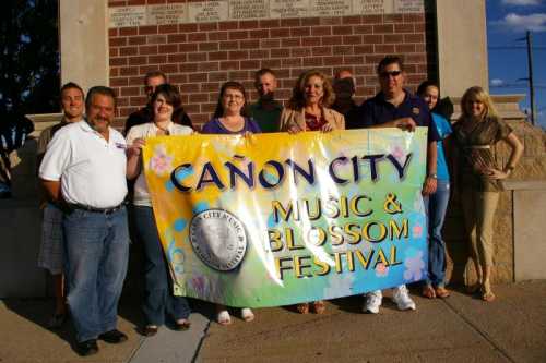 Group of people holding a banner for the Canon City Music & Blossom Festival, smiling outdoors.