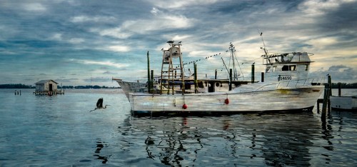 A weathered fishing boat docked in calm waters, with a bird flying nearby under a cloudy sky.