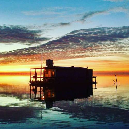A serene sunset over a calm lake, featuring a small houseboat reflecting on the water's surface.