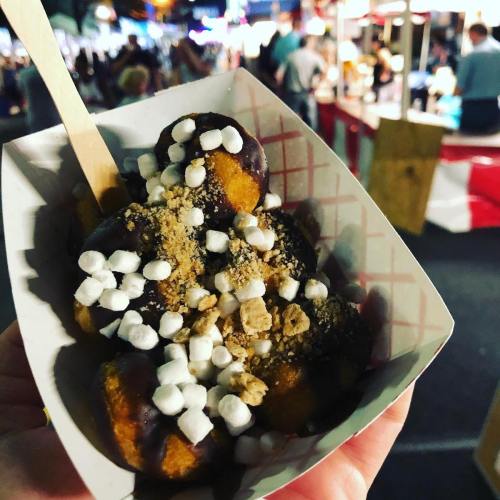 A hand holding a tray of dessert balls topped with chocolate, crushed graham crackers, and mini marshmallows at a fair.