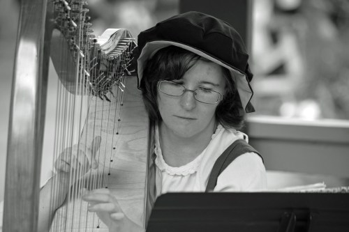 A person in period clothing plays a harp, focused and wearing glasses, in a softly lit setting.