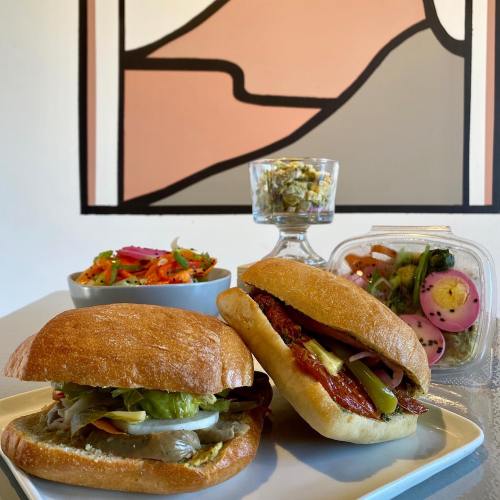 Two sandwiches with toppings on a plate, accompanied by a colorful salad and a dessert in a glass, with a mural in the background.
