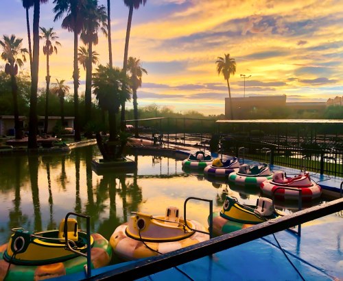 Colorful bumper boats sit in still water at sunset, with palm trees silhouetted against a vibrant sky.