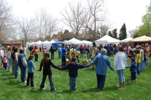 A diverse group of people holding hands in a circle on a grassy field, surrounded by tents and trees.